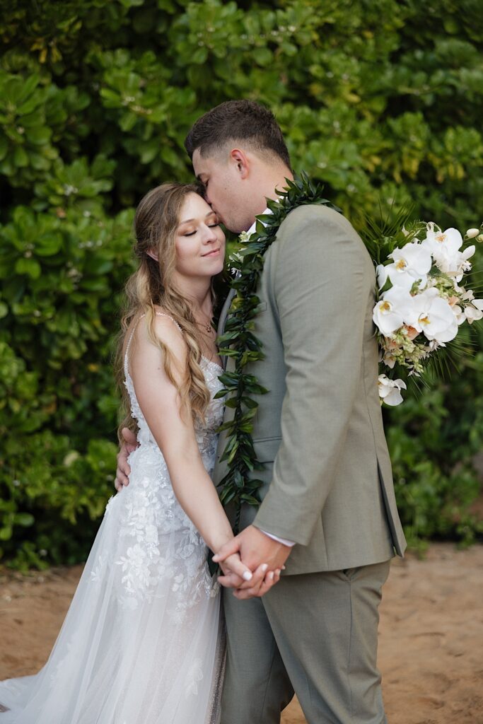 Bride in boho style dress, holding tropical flower arrangement leans into Groom as he kisses her forehead during their elopement ceremony in Maui, Hawaii. 