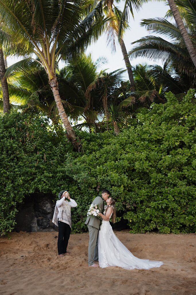 Bride in white Boho style dress kisses groom in grey suit while officiant stands next to them as during their Maui Elopement in Hawaii. 