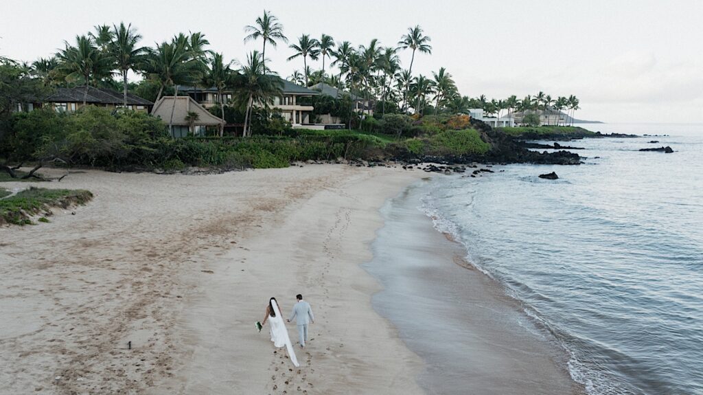 Hawaiian Elopement Photographer captures bride and groom from far away walking along the beach front towards beautiful beach houses after their Maui Elopement Wedding Ceremony.  