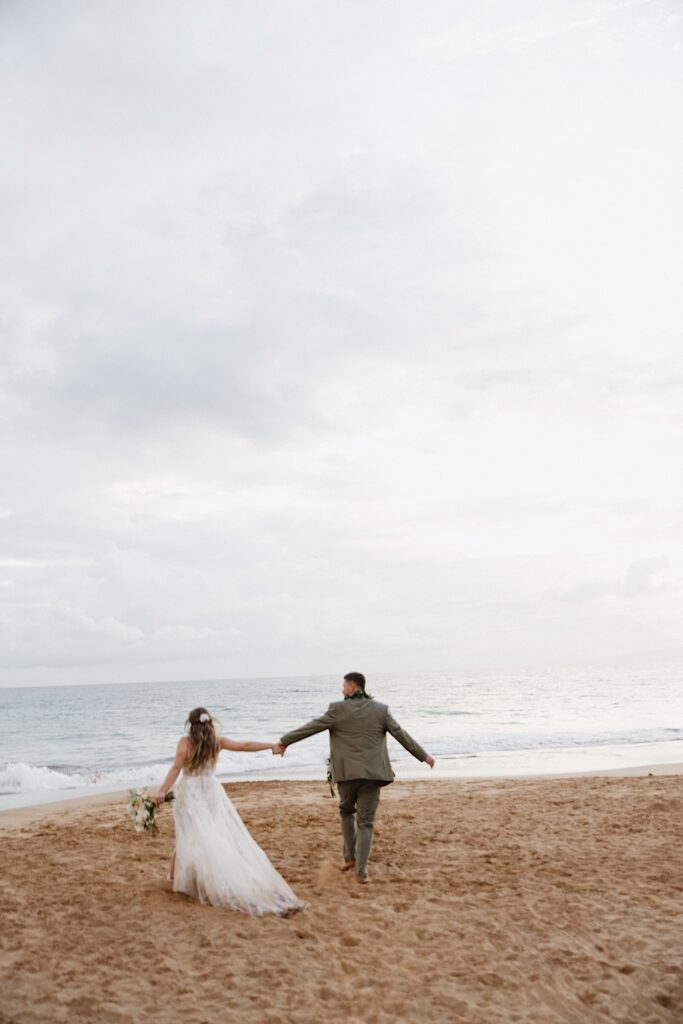 Bride in Boho style dress holds hands with Groom in green suit as they run into the Ocean after their elopement wedding ceremony in Maui Hawaii. 