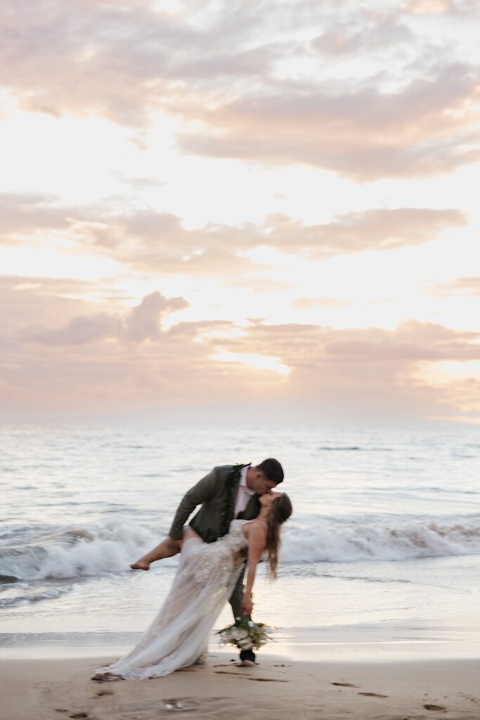 Groom in green suit dips bride in beautiful Boho style wedding dress on sandy beach after elopement wedding ceremony in Maui Hawaii. 