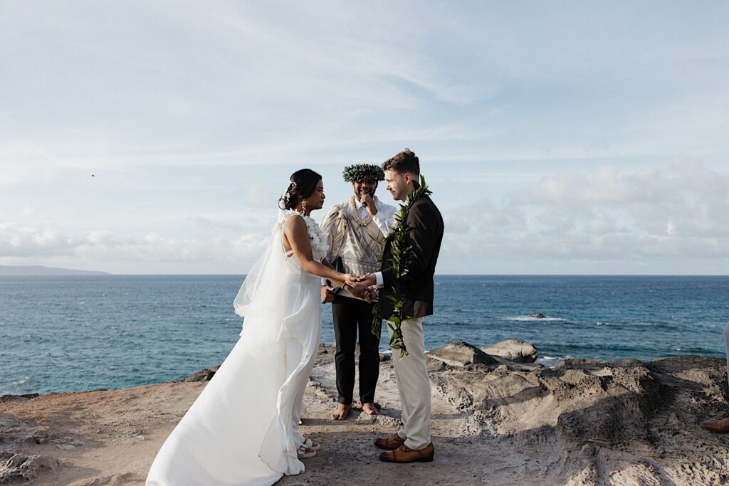 Bride in beautiful white dress and veil holds hands with Groom in Brown suit jacket and Khaki pants as their officiant marries them on beautiful cliff overlooking the Ocean during Maui Elopement Wedding ceremony. 