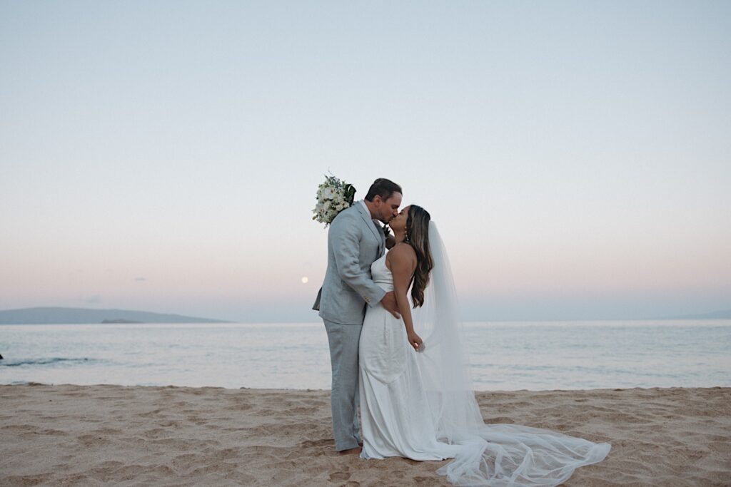 Bride in white silk dress kisses groom in grey wedding suit on the beach during Maui Elopement Wedding Ceremony. 