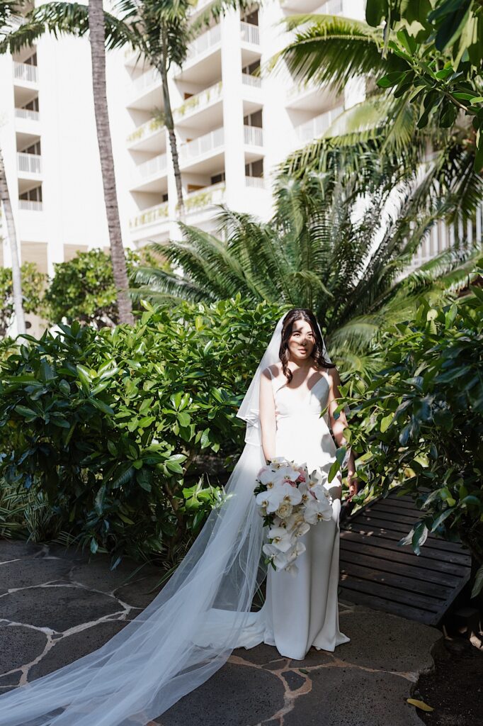 Bride with a long white veil stands under shade of palm trees and tropical foliage at the beautiful Four Seasons Hotel in Oahu Hawaii 