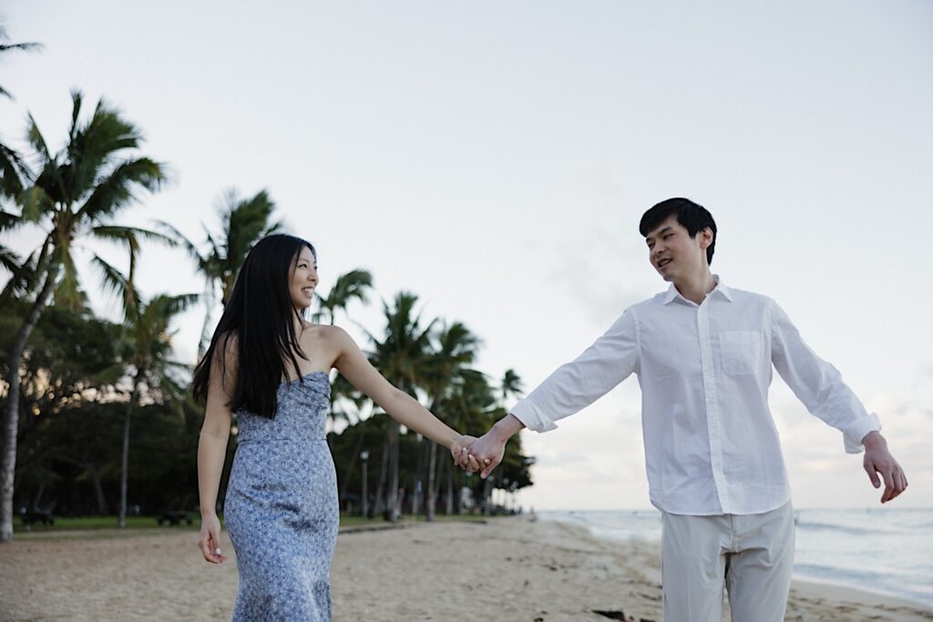 Girl in blue floral dress holds hand with her husband in a white shirt while walking across a palm tree lined beach front on a beautiful Hawaiian beach resort. 