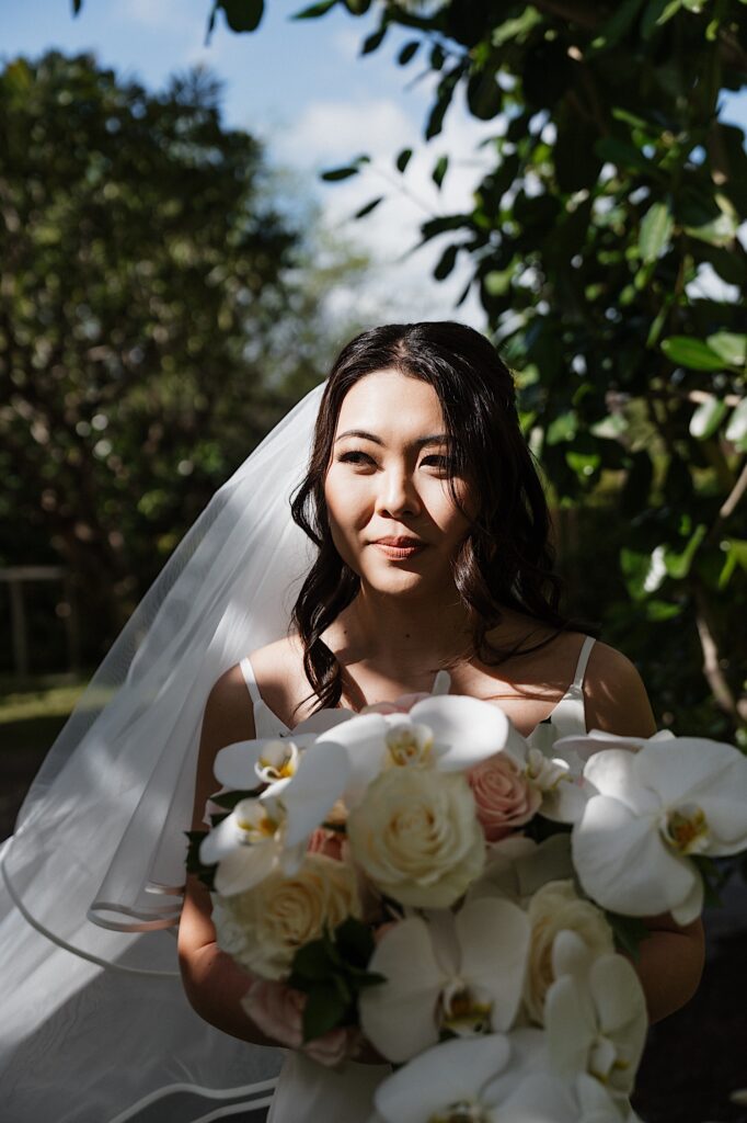 Bride shows beautiful tropical bouquet standing under trees at before ceremony at the Four Seasons in Oahu Hawaii. 