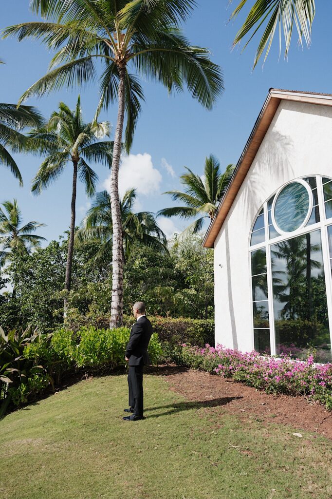 Groom in black tux stands with his back turned as he waits for the first look with his bride near a chapel before intimate wedding ceremony at Four Seasons Oahu in Hawaii. 