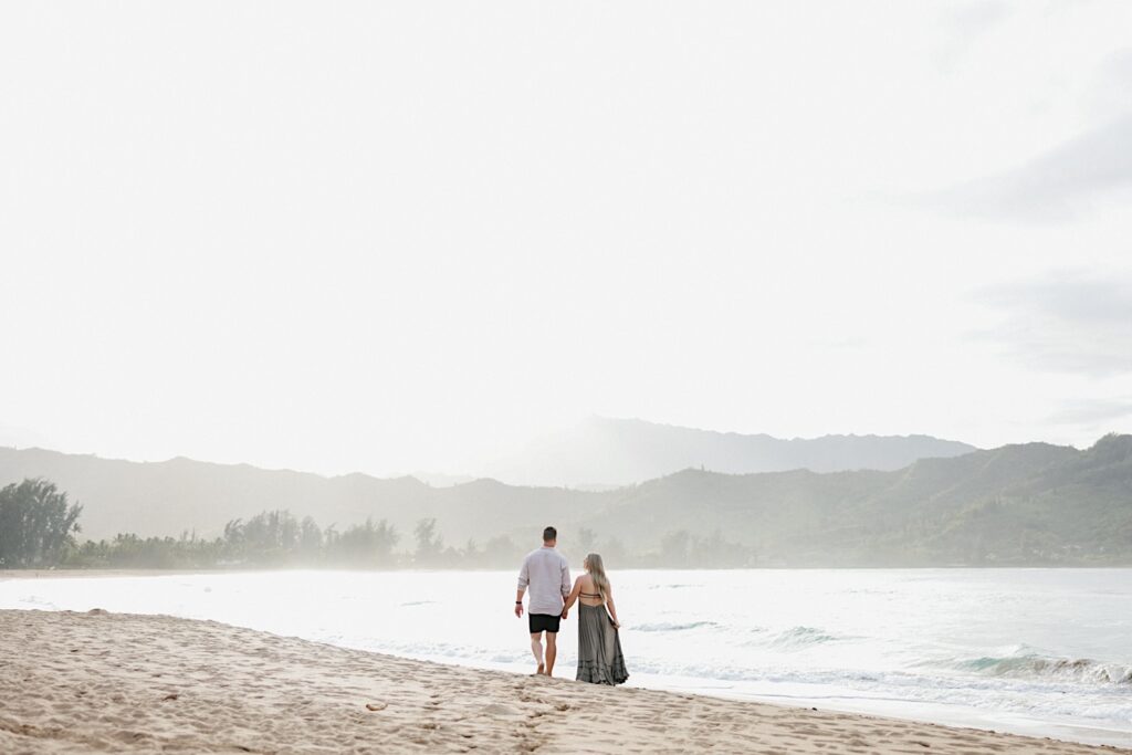 Couple holds hands walking along the beach with a mountain-scape background on a resort vacation in Hawaii.