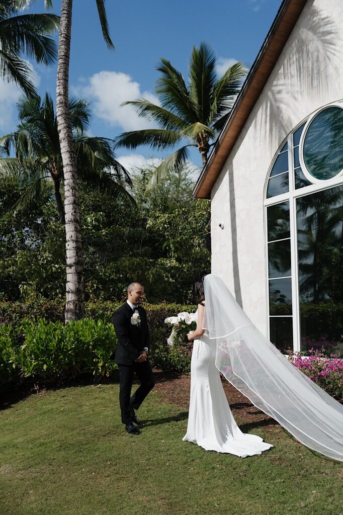 Groom in black tux smiles seeing bride for the first time outside beautiful white chapel surrounded by palm trees before intimate wedding ceremony at Four Seasons Oahu in Hawaii. 