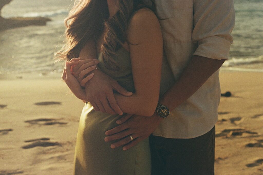 Hawaii Engagement photographer captures a couple hugging on a beautiful beach front on a vacation resort perfect for couples in Hawaii. The man is wearing a white shirt with a big watch on his wrist and his wife is wearing a beautiful silky sage dress. 