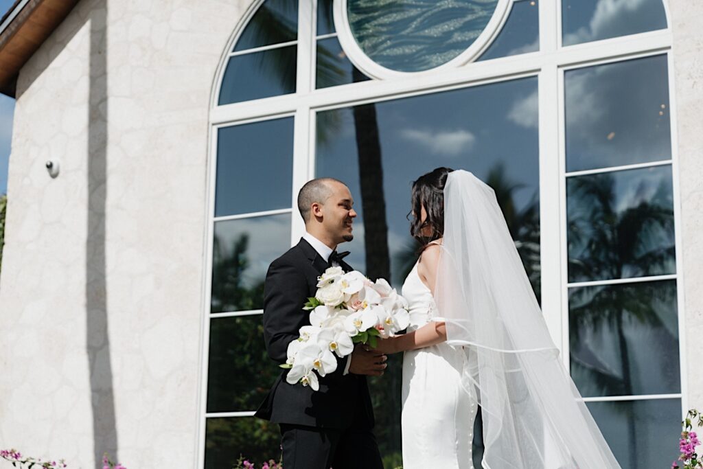 Bride and Groom look into each other's eyes standing in front of large Chapel window at the Four Seasons Oahu in Hawaii. 