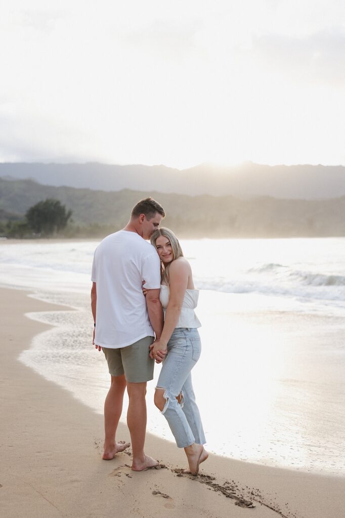 Couple in white romantically stand together on a beach front with a mountain backdrop at a beautiful Hawaiian resort for couples. 