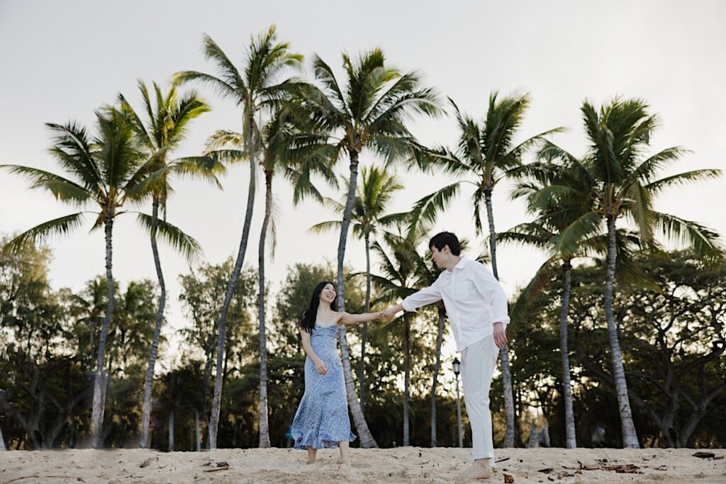 A woman in a blue floral dress dances with her husband in a white shirt and khaki pants on sandy beach with a palm tree background at a Hawaiian resort for couples. 