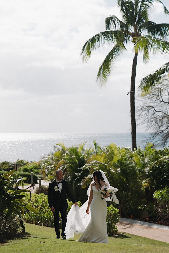 Groom Holds the train of bride's long white veil as they walk through the grass surrounded by tropical plants at Four Seasons Oahu in Hawaii. 