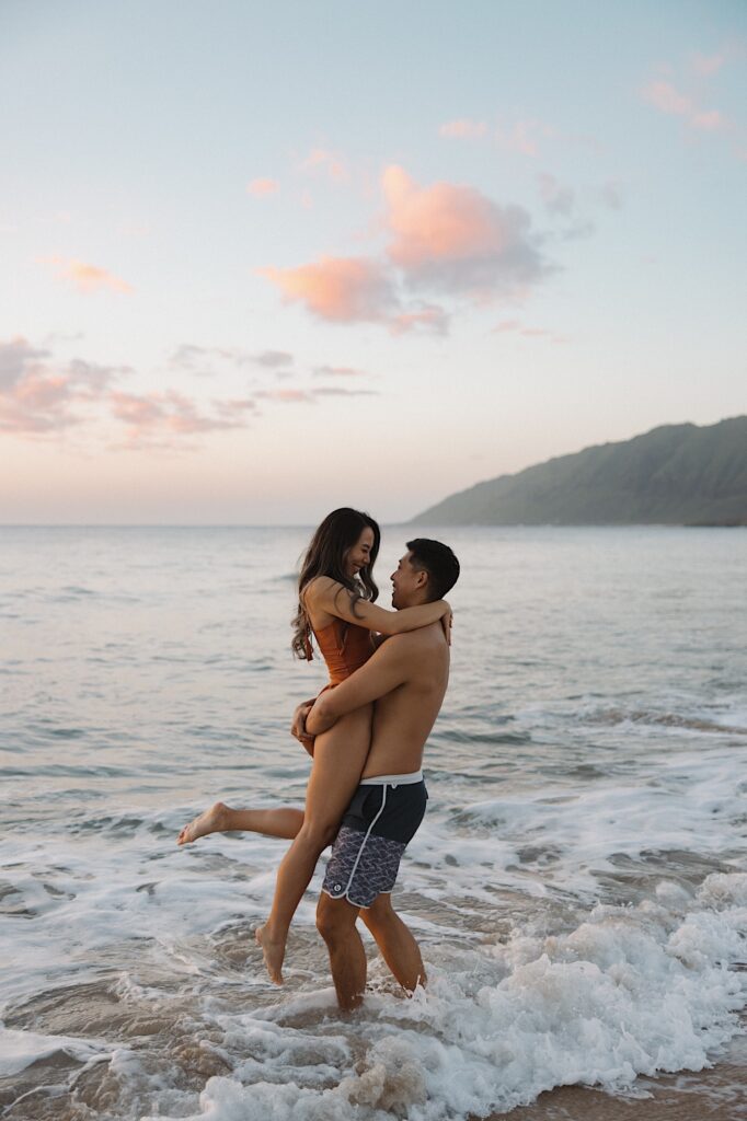 Husband in blue board shorts lifts up his wife in a orange bathing suit in the waves at a Hawaiian beach resort with a mountain scape background. 