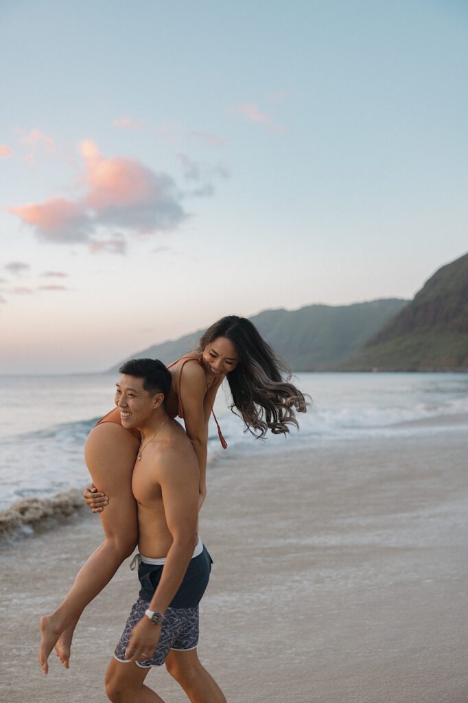 Husband in blue board shorts carries his wife in an orange bathing suit on a beautiful Hawaiian beach resort on vacation. 