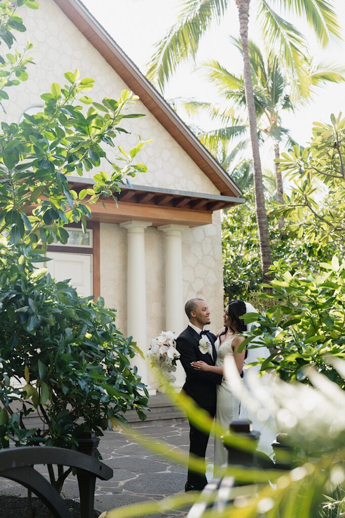 Bride and Groom hold each other outside chapel surrounded by tropical  plants at the Four Seasons Oahu in Hawaii. 