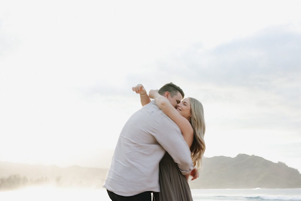 Couple hugs during engagement photos with the ocean in the background with mountains in the background at a Hawaiian vacation resort. 