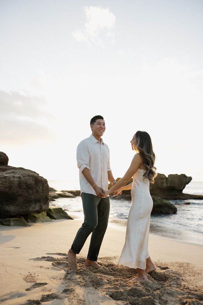 Man in white shirt and grey pants holds hands with his wife in a beautiful silky white dress on the beach at a Hawaiian resort for couples.