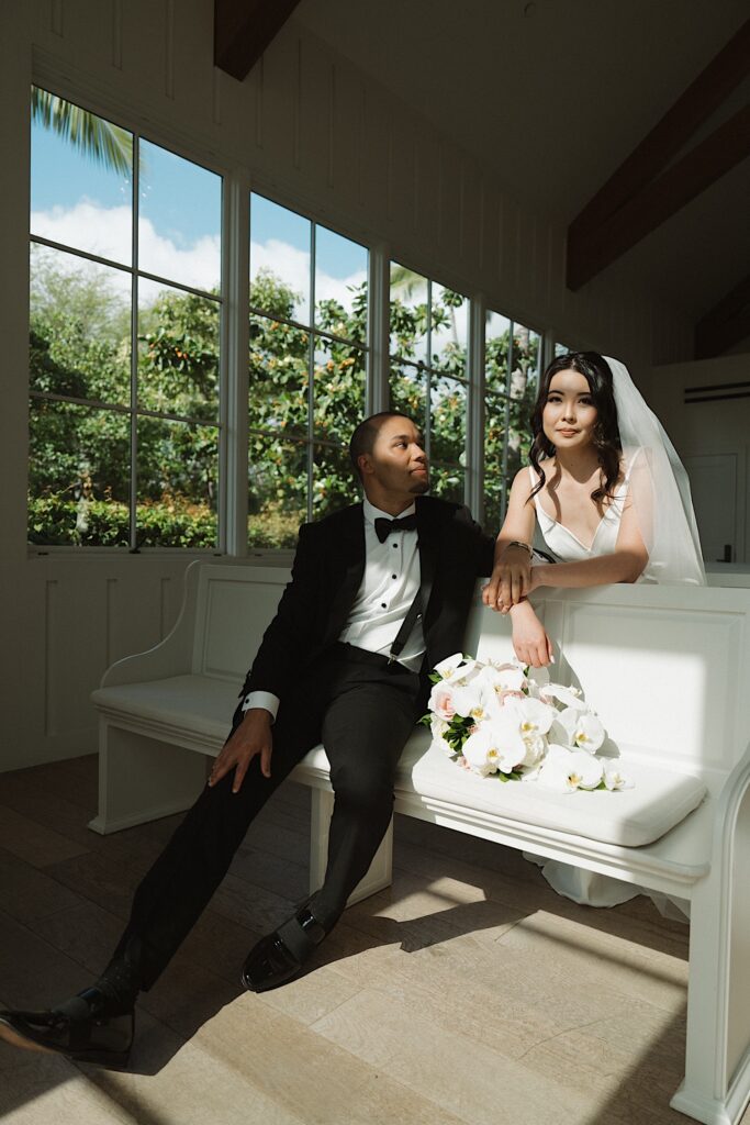 Bride leans over Chapel Pew at Four Seasons Oahu in Hawaii, looking into the camera next to Groom who is sitting on the pew looking at the bride. 