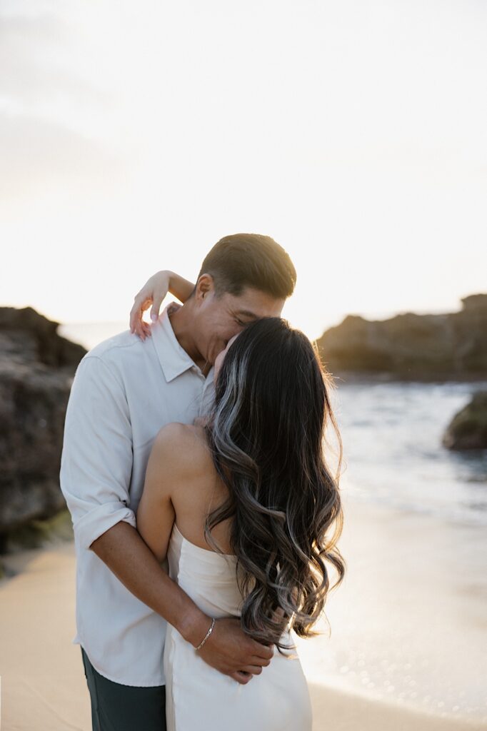 Man in a white shirt kisses a woman in white silky dress on a beautiful Hawaiian beach at sunset. 