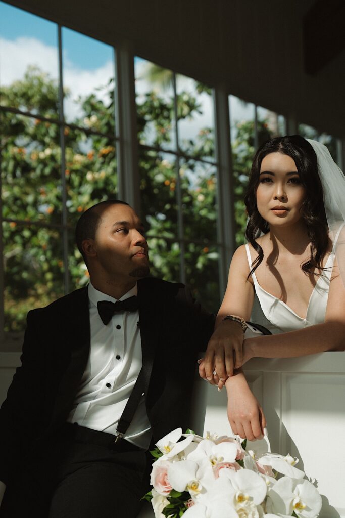 Bride leans over Chapel Pew at Four Seasons Oahu in Hawaii, looking into the camera next to Groom who is sitting on the pew looking at the bride. 