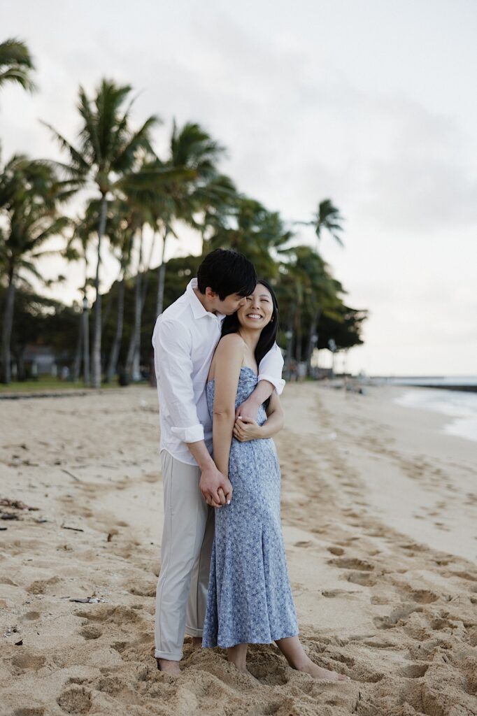 Man in a white shirt and khaki pants hugs a woman in a blue floral sundress from behind on a beach with palm trees in the background at a Hawaiian resort for couples. 