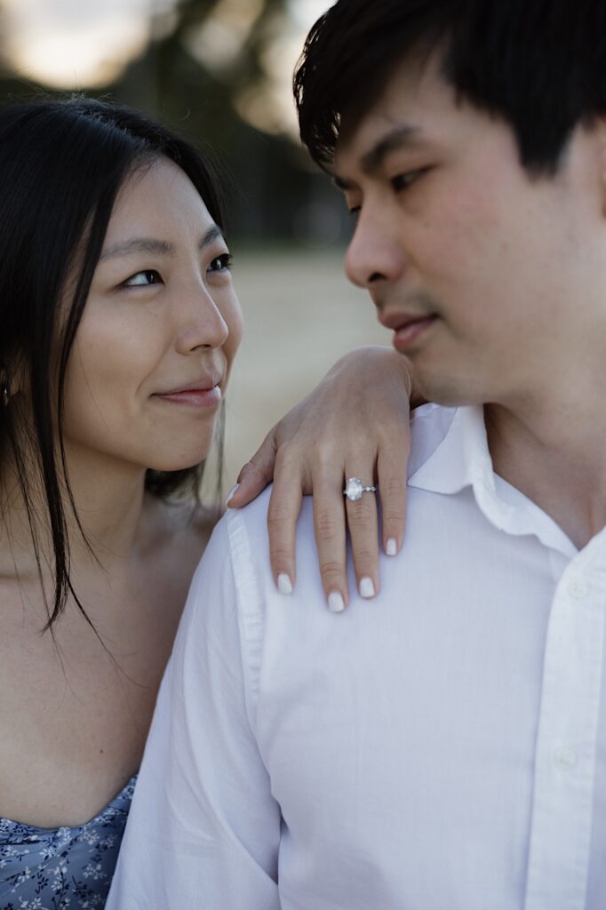 Hawaii Engagement photographer captures a woman showing off her engagement ring with her hand on her husband's shoulder on a beautiful beach at a Hawaiian resort.  