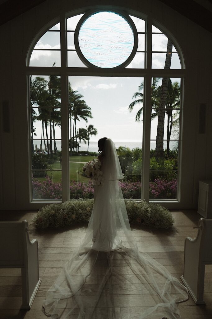 Bride stands in front of brightly lit window showing palm trees and beach front in her beautiful gown and veil before wedding ceremony at Four Seasons Oahu in Hawaii. 