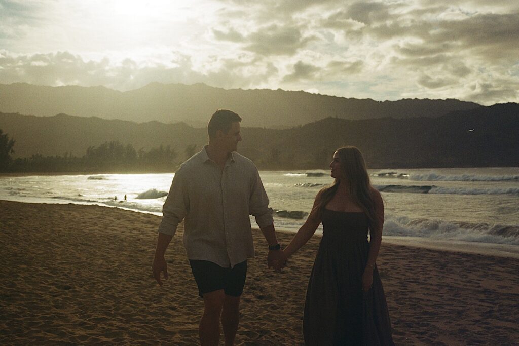 Man in a white button up holds hand with his wife in a blue dress walking along the beach at dusk with the ocean in the background. 