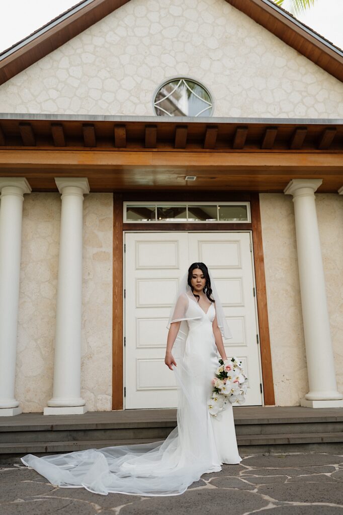 Bride poses in her beautiful white gown and long white veil holding tropical bouquet in front of white chapel at Four Seasons Oahu in Hawaii. 