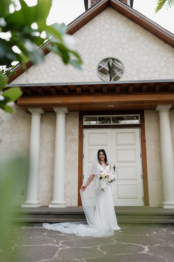 Bride poses smiling in her beautiful white gown and holding her long white veil holding tropical bouquet in front of white chapel at Four Seasons Oahu in Hawaii. 