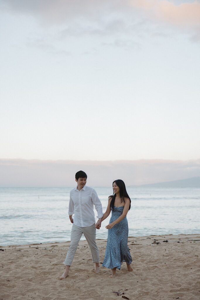 Man in white shirt with khaki pants holds hands with a woman in a blue floral dress on a beautiful beach with the ocean in the background at a Hawaiian vacation resort for couples.