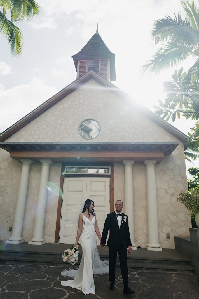 Bride in white flowy dress walks with her groom in a black tux and black bow tie outside of chapel at Four Seasons Oahu in Hawaii. 