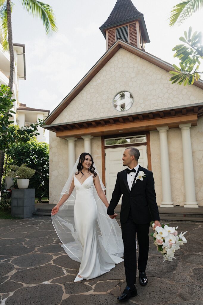 Bride in white flowy dress walks smiling with her groom in a black tux and black bow tie outside of chapel at Four Seasons Oahu in Hawaii. 