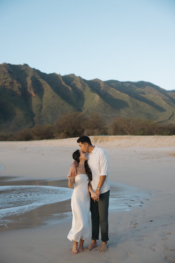 Man in white shirt kisses a woman in a silky white dress on the forehead on the beach with an ocean and mountain backdrop at a resort vacation spot for couples in Hawaii. 