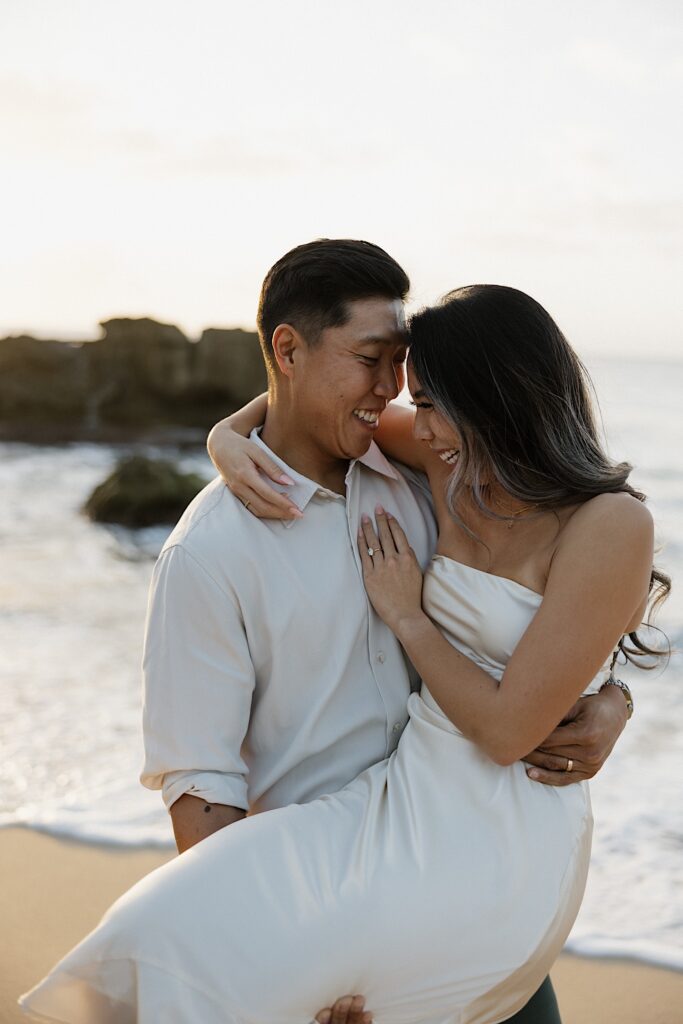 Man in white bottom up carries a woman in a white silky dress as they laugh together on the beach with an ocean background at a couples resort in Hawaii. 