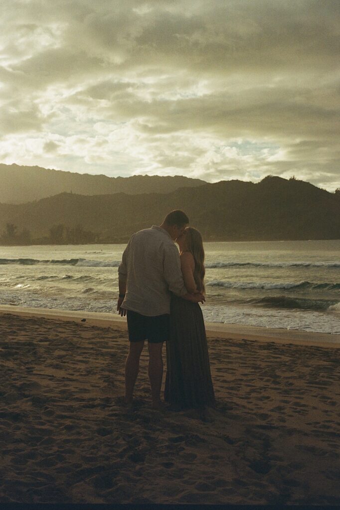 Couple kisses at dusk with the ocean and mountains in the background at a beautiful couples resort in Hawaii. 