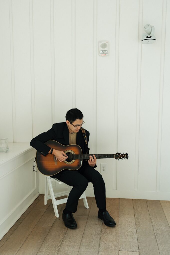 Male Guitarist sits on a white chair playing live music inside Four Seasons Oahu in Hawaii during intimate wedding ceremony. 