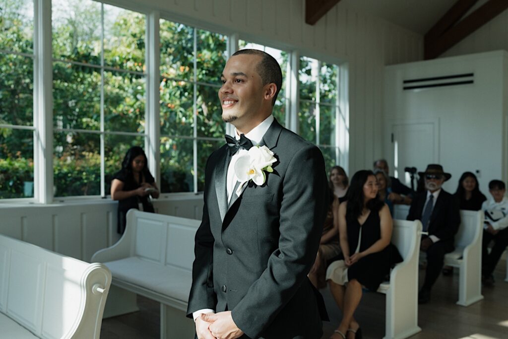 Groom in a black tux with black bow tie walks down the aisle smiling during intimate wedding ceremony in Four Seasons Oahu chapel in Hawaii. 