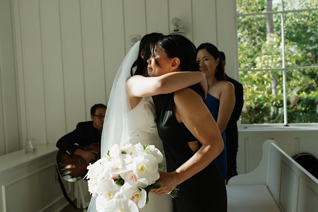 Bride hugs her mom holding beautiful tropical white bouquet during intimate ceremony at Four Seasons in Oahu Hawaii. 