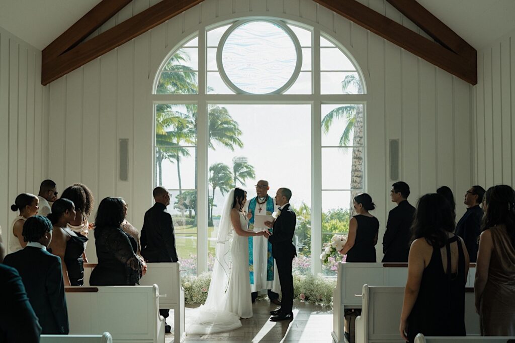 Bride and groom hold hands at the end of the aisle in front of beautiful chapel window looking out on tropical flowers and trees. 