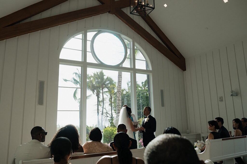 Bride and groom hold hands at the end of the aisle in front of beautiful chapel window looking out on tropical flowers and trees. 