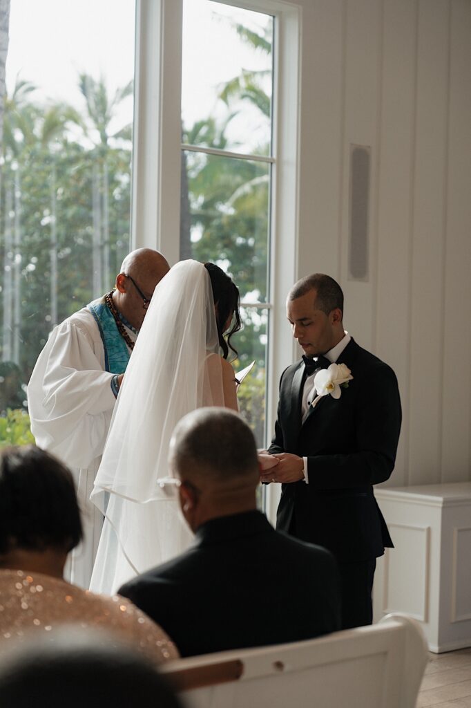 Groom places ring on her finger during intimate wedding ceremony at Four Seasons Oahu Chapel in Hawaii.