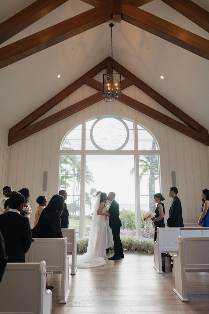 Bride and Groom kiss at the end of the aisle in front of beautiful scenic window in intimate chapel at Four Seasons Oahu in Hawaii