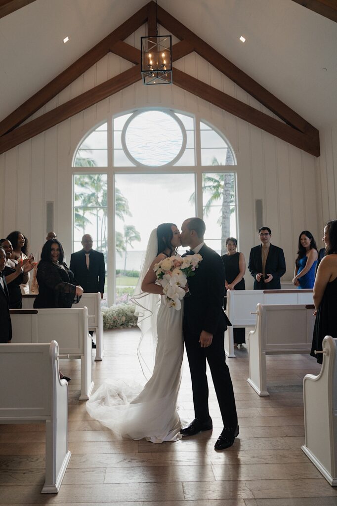 Bride and Groom kiss at the end of the aisle as guests smile in front of beautiful scenic window in intimate chapel at Four Seasons Oahu in Hawaii