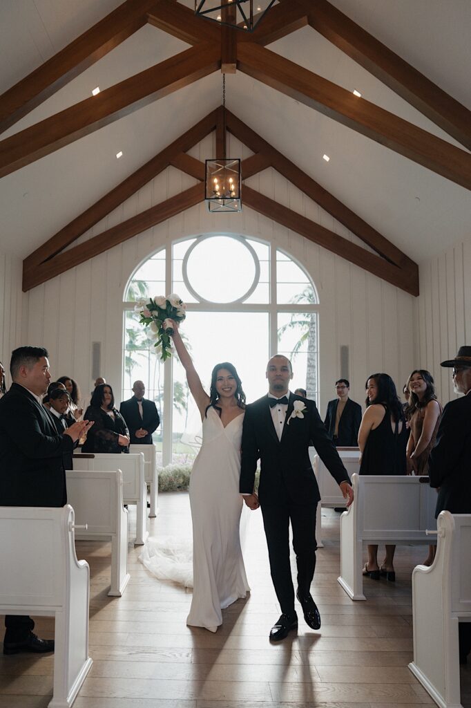 Bride with her arm raised holding beautiful tropical bouquet holding hands with the groom as they walk down the aisle in intimate chapel at Four Seasons Oahu in Hawaii.