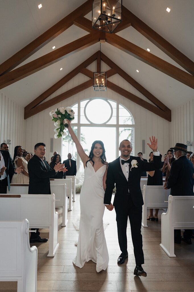 Bride with her arm raised holding beautiful tropical bouquet holding hands with the groom as they walk down the aisle in intimate chapel at Four Seasons Oahu in Hawaii.