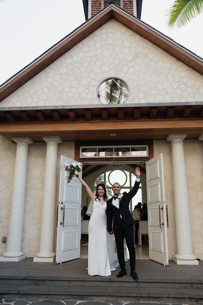 Bride with her arm raised holding beautiful tropical bouquet holding hands with the groom outside intimate chapel at Four Seasons Oahu in Hawaii.