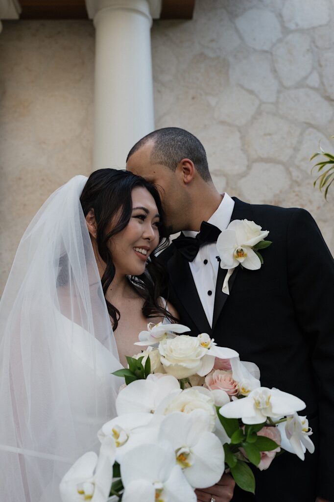 Groom whispers something into the bride's ear as she smiles brightly holding her tropical bouquet outside Four Seasons Oahu Wedding Chapel in Hawaii. 